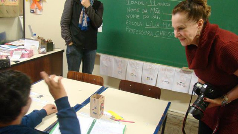 Primeira-Dama do Estado Sandra Genro, segurando a máquina fotográfica, reclinada, conversando em Libras com um aluno da Escola Lilia Mazeron, com tradução simultânea da professora.