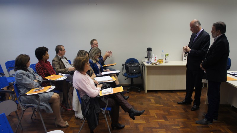 Descrição da imagem: Prefeitos e representantes dos municípios da Associação dos Municípios do Vale do Rio dos Sinos (AMVRS) estão sentados na sala de reuniões localizada no prédio lilás da Feevale, e de pé o presidente da Faders Acessibilidade e