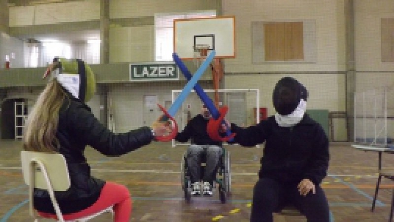Descrição da imagem: Ginásio de esportes e no centro do local, o atleta paralímpico de Esgrima em Cadeira de Rodas, Maurício Stempniak de Lima, demonstrando com dois alunos a vivência no esporte paralímpico.Crédito: Divulgação Fundergs
