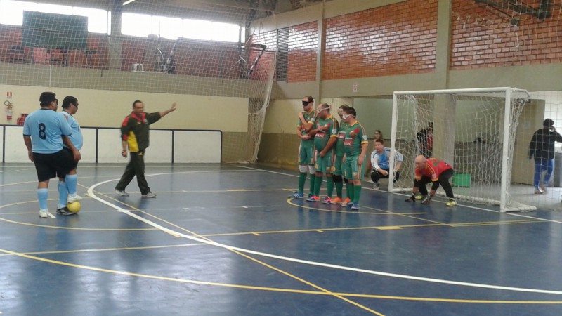 Descrição da imagem: Ginásio de esportes com quadra de futebol, estando dois jogadores de uniformes azuis se posicionando para cobrar falta e outros quatro de verde formam barreira em frente a goleira, onde também está posicionado o goleiro. No lado