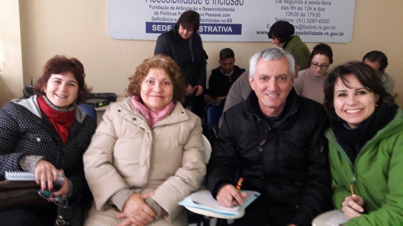 Descrição: Grupo de quatro pessoas, em uma sala de curso, três mulheres e um homem em plano americano; todos adultos de pele clara; vestindo roupas de inverno, todos com expressão sorridente, sentados em cadeiras móveis escolares estofadas na cor azul e