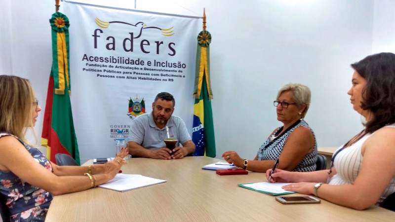 Quatro pessoas sentadas ao redor da mesa, estão em reunião no gabinete da presidência da FADERS. Ao fundo da imagem, banner da FADERS, ao lado esquerdo do banner, bandeira do Rio Grande do Sul, ao lado direito bandeira do Brasil.