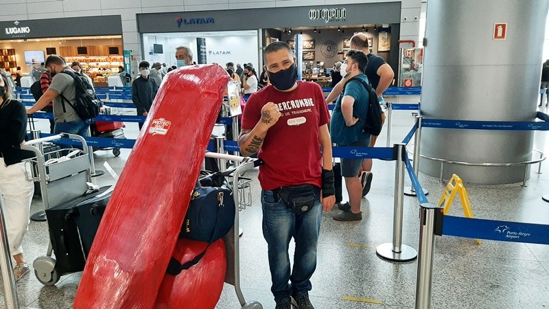 Um homem usando máscara, com deficiência no braço esquerdo, posa para a foto mostrando o punho direito. À esquerda, um carrinho com bagagens. Ao fundo, fitas azuis que demarcam as filas do aeroporto e pessoas com malas e mochilas.