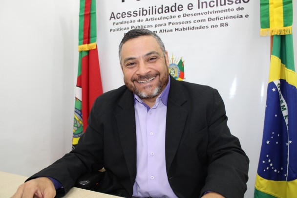 O presidente da FADERS, Marquinho Lang, posa para a foto. Ele usa camisa, paletó, e está sorrindo. Atrás dele, o banner da FADERS entre as bandeiras do Rio Grande do Sul e do Brasil.