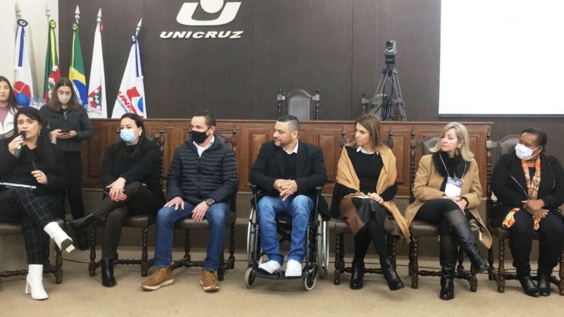 Sete pessoas, cinco mulheres e dois homens, estão sentados em frente a um painel com a logomarca da Unicruz e de cinco bandeiras, entre elas a do RS, do Brasil, de Cruz Alta e da Unicruz. Seis pessoas olham para a mulher que está a direita falando ao microfone.