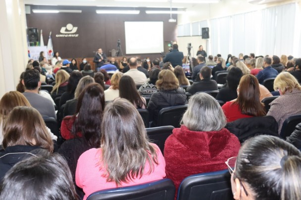 Foto tirada do fundo do auditório. Dezenas de pessoas estão de costas observando um homem que fala em pé ao microfone. Atrás dele, as pessoas que compuseram a mesa. Atrás, um banner da FADERS Acessibilidade e Inclusão e, na parede, a logomarca Unicruz.