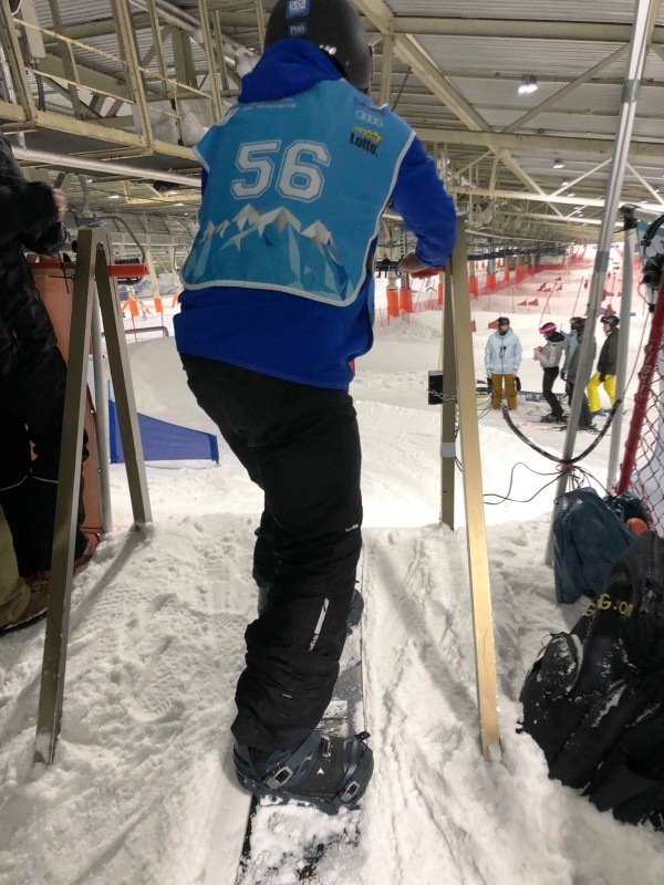 De costas, o para atleta está em pé sobre uma prancha sobre a neve, na posição de largada. Ele está com um colete azul com o número 56. Ao fundo, bandeiras vermelhas que indicam o percurso a ser percorrido.