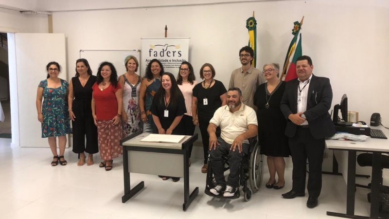 Doze pessoas, nove mulheres e três homens, posam para a foto. Todos estão em pé, exceto o presidente da FADERS,  Marquinho Lang, que está sentado sobre cadeira de rodas. Ao fundo, o banner da FADERS e as bandeiras do Brasil e do Rio Grande do Sul.