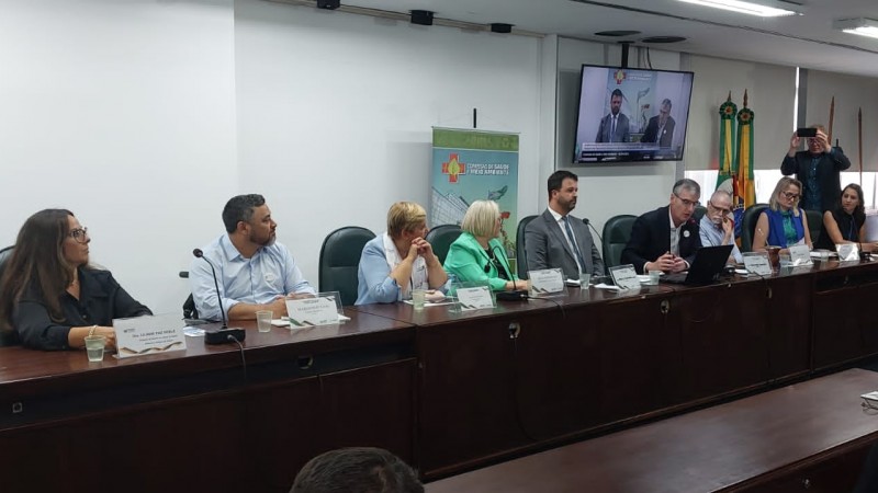 Nove pessoas, seis mulheres e três homens, estão sentados atrás de uma mesa. Marquinho Lang, presidente da FADERS, é o segundo da esquerda para direita. Ao fundo, o banner da Comissão de Saúde da Assembleia Legislativa, e uma televisão pendurada na parede, transmitindo a reunião ao vivo. Um homem está em pé, à direita, atrás dos integrantes da mesa, fazendo uma foto pelo celular.