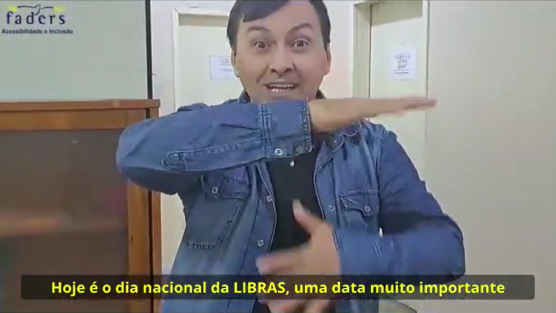 Frame do vídeo institucional. Um homem está fazendo sinais em Libras. Na legenda, está escrito "Hoje é o dia nacional da Libras, uma data muito importante". No alto, à esquerda, a logomarca da FADERS.