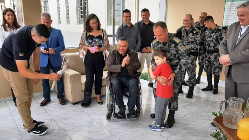 Um menino, usuário da Equoterapia, abraça o Comandante de Polícia de Choque, coronel Claudio Azevedo Goggia. à esquerda, está o presidente da FADER, Marquinho Lang, sentado em cadeira de rodas, aplaudindo . Ao redor, outras doze pessoas: duas mulheres, Alane e Eva; quatro homens com uniformes da BM; e cinco homens à paisana.