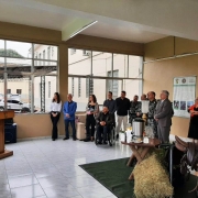 À esquerda, o Comandante de Polícia de Choque, coronel Claudio Azevedo Goggia, fala ao microfone, posicionado atrás de um púlpito de madeira. À direita, observam-no o presidente da FADERS, Marquinho Lang, sentado sobre cadeira de rodas; quatro mulheres, cinco homens fardados com uniforme da Brigada Militar; e outros quatro homens em trajes civis.