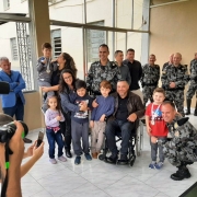 Posam para a foto quatro crianças em pé ao redor do presidente da FADERs, Marquinho Lang, que está sentado em cadeira de rodas, o Comandante de Polícia de Choque, coronel Claudio Azevedo Goggia, agachado, o comandante do 4º RPMon, Ives Cláudio Pacheco, em pé atrás do grupo, uma mãe que abraça um menino que está ao lado de Lang, e outra mãe, que segura outro menino no colo. Ao redor, civis e militares observam a cena. 