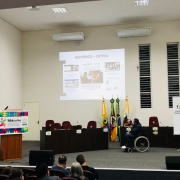 Sentado em cadeira de rodas, Marquinho Lang fala ao microfone enquanto aponta para uma imagem projetada na parede com o histórico da Ciptea. Ele é observado por Aline Machado, em pé, à esquerda. A frente do presidente da FADERS, o público observa, sentado.