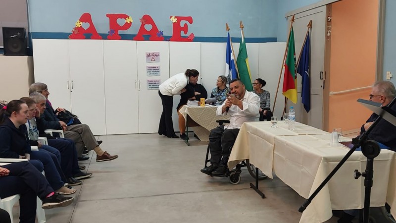 Sentado sobre cadeira de rodas, Marquinho Lang fala ao microfone. À direita, sentado atrás de uma mesa, um homem. À esquerda, o público ouve atentamente. Atrás de Lang, quatro mulheres estão ao redor de uma mesa. Na parede, em vermelho, está escrito APAE.