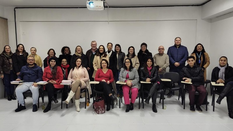 Vinte e quatro pessoas, nove delas sentadas em cadeiras com apoio para escrever e o restante em pé, posam para a foto dentro da sala de aula.