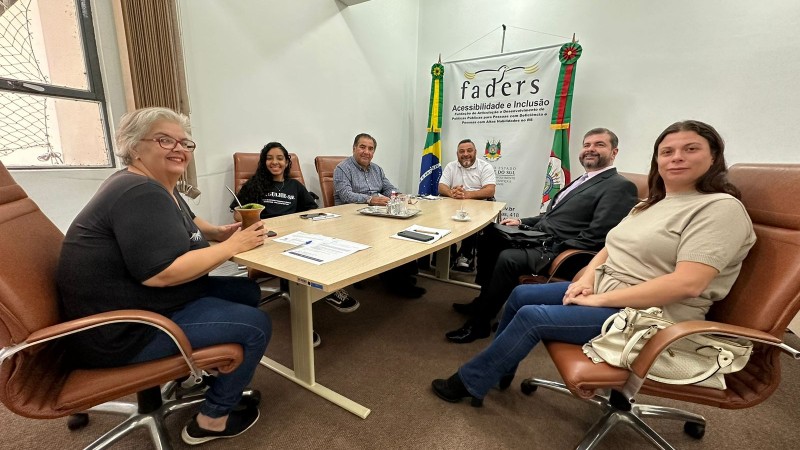 Sala com paredes brancas e carpete marrom, ao centro mesa grande e bege com cadeiras em couro em marrom, onde estão o presidente da Faders e a diretora técnica nas pontas e ao redor os representantes da Secretaria de Justiça RS, ao fundo na parede o Banner da Faders.