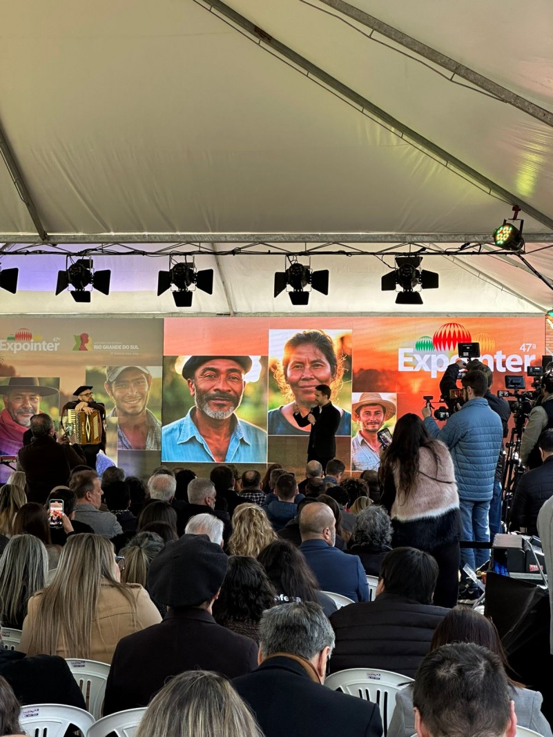 foto - Governador Eduardo Leite faz seu pronunciamento no lançamento da 47ª Expointer. no palco ele esta a direita, no fundo um painel com foto de trabalhadores rurais e a logo da Expointer, o publico esta sentado em cadeiras de plástico brancas
