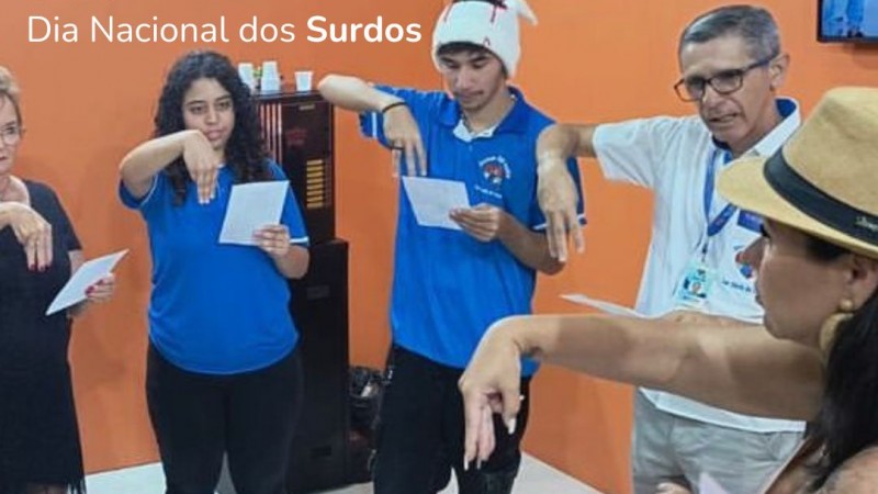 Card Dia Nacional do Surdo - ilsutrando no fundo uma foto da professora de Libras da Faders Patrícia ensinando a Língua Brasileira de Sinais para pessoas durante evento em Caxias do Sul, aparece ela realizando um sinal e os alunos em sua frente também fazendo o mesmo sinal.... as paredes ao fundo são laranjas, e o chão é branco... no canto esquerdo do card em branco tem a escrita - 26.09 DIA NACIONAL DO SURDO, e no final do card centralizado tem escrito em branco - SINAIS SÃO PARA OS OLHOS O QUE AS PALAVRAS SÃO PARA OS OUVIDOS!, abaixo a logo da Faders e ao lado direito e esquerdo da logo há imagens de sinais em transparência azul. Fim da descrição