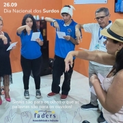 Card Dia Nacional do Surdo - ilsutrando no fundo uma foto da professora de Libras da Faders Patrícia ensinando a Língua Brasileira de Sinais para pessoas durante evento em Caxias do Sul, aparece ela realizando um sinal e os alunos em sua frente também fazendo o mesmo sinal.... as paredes ao fundo são laranjas, e o chão é branco... no canto esquerdo do card em branco tem a escrita - 26.09 DIA NACIONAL DO SURDO, e no final do card centralizado tem escrito em branco - SINAIS SÃO PARA OS OLHOS O QUE AS PALAVRAS SÃO PARA OS OUVIDOS!, abaixo a logo da Faders e ao lado direito e esquerdo da logo há imagens de sinais em transparência azul. Fim da descrição