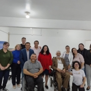 Foto: em suas cadeiras de rodas os presidentes da Faders e do Coepede, em pé os participantes da inauguração da nova sede do Conselho que aconteceu neste dia 01 pela manhã e fica no predio da Faders na Morretes. Ao fundo paredes brancas, teto branco, e piso branco. Fim da descrição