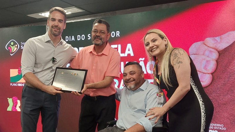 Foto- no palco do Auditório do Caff onde aconteceu a solenidade de entrega do prêmio, o seu Edison e sua esposa posam para foto com a placa de reconhecimento entregue pelo Governador Eduardo Leite e ao lado o presidente da Faders, Marco Antonio Lang e a secretária de Planejamento do RS. No fundo um painel com as logos do Governdo Estado da Semana do Servidor da Secretaria de Planejamento, e com a logo do Premio.  Fim da Descrição