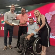 Foto- no palco do Auditório do Caff onde aconteceu a solenidade de entrega do prêmio, o seu Edison e sua esposa posam para foto com a placa de reconhecimento entregue pelo Governador Eduardo Leite e ao lado o presidente da Faders, Marco Antonio Lang e a secretária de Planejamento do RS. No fundo um painel com as logos do Governdo Estado da Semana do Servidor da Secretaria de Planejamento, e com a logo do Premio.  Fim da Descrição