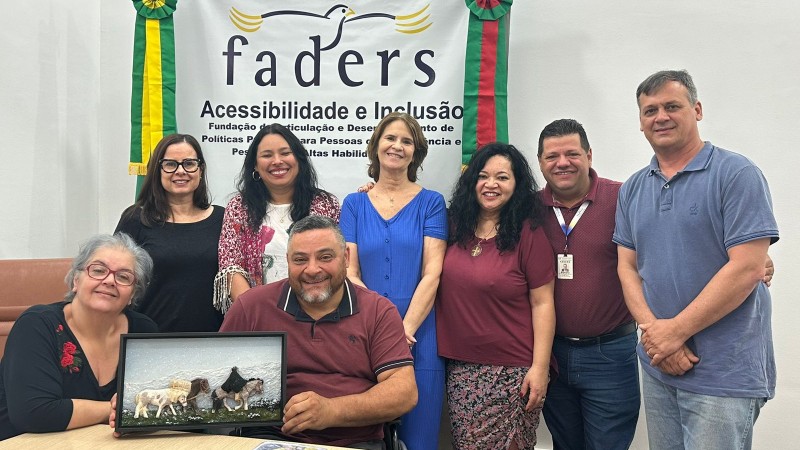 Foto – da esquerda para direita- sentada em uma cadeira marrom e em sua frente uma mesa em madeira clara a Diretora Técnica da Faders segura a prancha tátil junto com o presidente da entidade que está ao seu lado em sua cadeira de rodas, em pé atrás deles a Coordenadora de Atendimento, a Coordenadora de Capacitação, a coordenadora de Acessibilidade, a coordenadora  de Direitos e Políticas Públicas, o diretor Administrativo e o Chefe de Gabinete da Faders.Ao fundo uma parede branca e o banner da faders,do lado direito dele a bandeira do RS e do esquerdo a do Brasil, no chão um carpete marrom. Fim da descrição.

