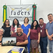 Foto – da esquerda para direita- sentada em uma cadeira marrom e em sua frente uma mesa em madeira clara a Diretora Técnica da Faders segura a prancha tátil junto com o presidente da entidade que está ao seu lado em sua cadeira de rodas, em pé atrás deles a Coordenadora de Atendimento, a Coordenadora de Capacitação, a coordenadora de Acessibilidade, a coordenadora  de Direitos e Políticas Públicas, o diretor Administrativo e o Chefe de Gabinete da Faders.Ao fundo uma parede branca e o banner da faders,do lado direito dele a bandeira do RS e do esquerdo a do Brasil, no chão um carpete marrom. Fim da descrição.

