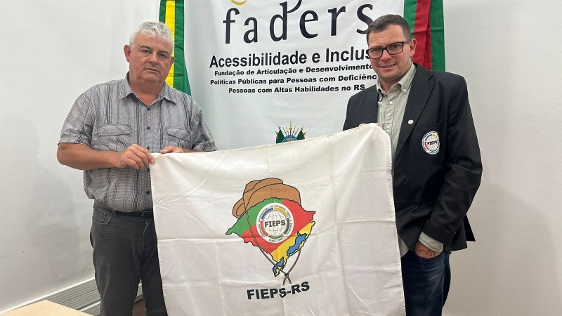 Foto - no fundo parede branca e o banner da Faders, a frente o servidor da Faders Paulo segura uma ponta da bandeira da Fieps-RS e na outra ponta o Delegado e Coordenador Deiques da Fiepes segura a bandeira, que é branca e possui no meio a logo da entidade. Fim da Descrição.