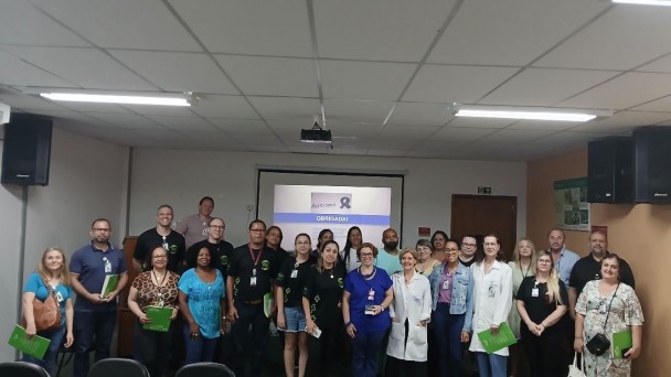 Foto do grupo que participou da palestra, ao centro a palestrante, ao fundo uma parede branca e uma tela com apresentação. Fim da descrição 