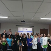 Foto do grupo que participou da palestra, ao centro a palestrante, ao fundo uma parede branca e uma tela com apresentação. Fim da descrição 