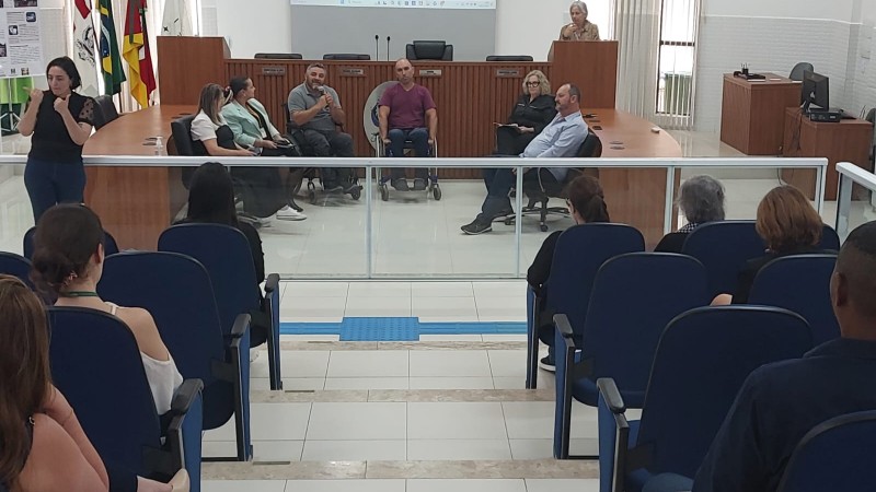 Foto: aparecem cadeiras azuis onde está o publico do evento, e na frente há uma separação em vidro, e então a mesa em formato de U em madeira, no fundo uma mesa mais alta ( inde fica a mesa diretora da Cãmara em dias de sessão) e há o pulpito, onde se pronuncia uma senhora loira de cabelos curtos e que veste bege. na Frente estão sentados os palestrantes em cadeiras pretas e o presidente da Faders em sua cadeira de rodas; Fim da descrição 