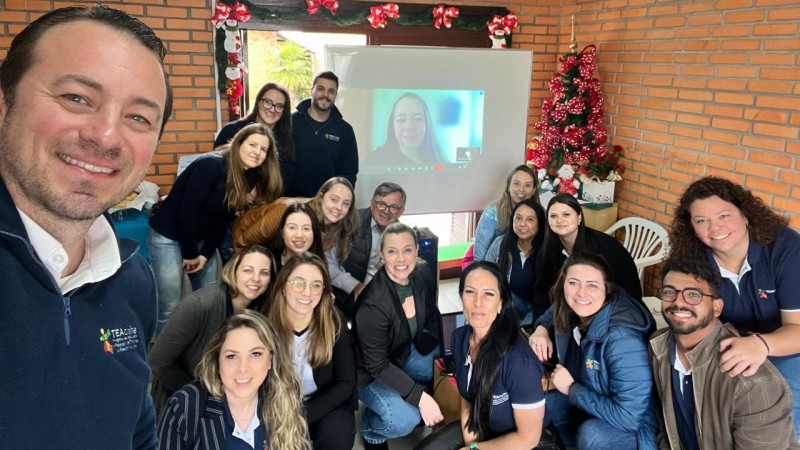 Foto- selfie, as paredes são em tijolo a vista, no fundo um painel onde aparece a coordenadora de Pesquisa da Faders On Line, e ao redor, uns em pé e uns abaixados, os profissionais que participaram da capacitação sobre Autismo e a Ciptea. Bem na frente quem fez a selfie é um homem claro, com cabelos curots castanhos, olho claro, e com uma blusa azul. Fim da Descrição