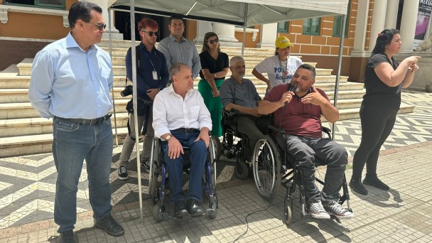 Na foto ao fundo o predio da Prefeitura de Porto Alegre, então em um toldo branco estão bem na frente, o presidente da Faders e sua cadeira de rodas com um microfone em mãos fazendo seu pronunciamento; ao seu lado a interprete de Libras da Faders, do outro o sec. Adjunto da Sec de Desenvolvimento Social de POA, e o vereador Alvoni Medina; atrás o jornalista que presentou o ceriomonial, ao seu lado o vereador Marcelo Bernardi, a representante da Defensoria Pública do RS, ao seu lado em sua cadeira de rodas o presidente do Coepede e a presidente do Comdepa em pé. Fim da Descrição 