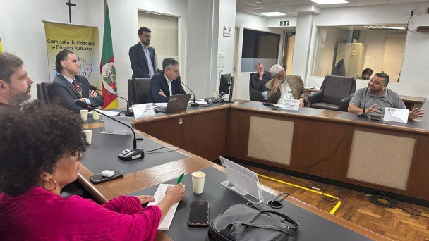 Foto - do espaço de convergência da Assembleia Legislativa onde aconteceu a audiência pública que debateu a Legai. No centro da sal uma mesa em formato de U, ela é em madeira com uns tampão em preto em cima e microfones pretos de mesa, as cadeiras aos seu redor saõ em couro pretas. Só na ponta do U há dois computadores, onde estão sentados o presidente da Frente Parlamentar em Defesa das Pessoas com Deficiencia da Asembleia e o secretario de Cidadania e Direitos humano do RS, ao redor da mesa estão os representantes de entidades que participaram dos debates, entre les o presidente da Faders. Fim da descrição