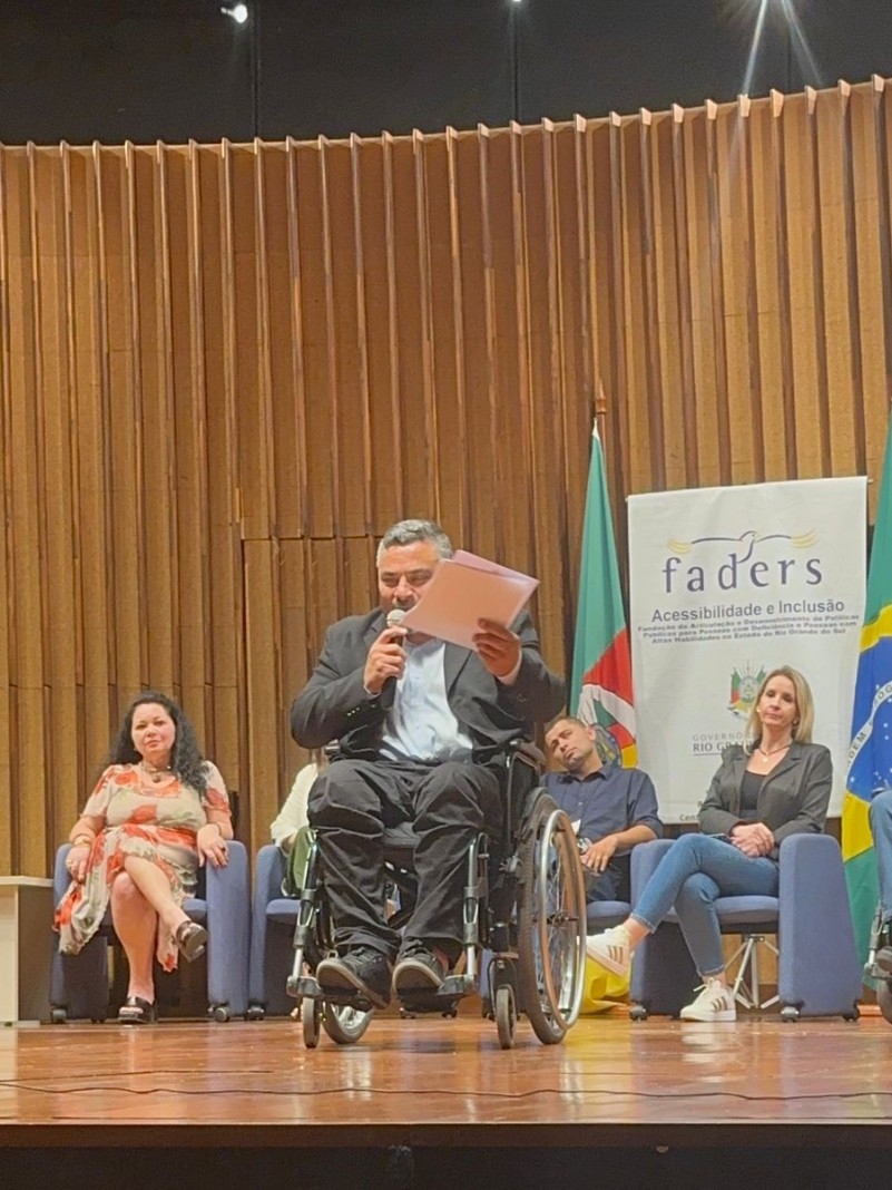 foto- no palco ao fundo o banner da faders, em cada lado uma bandeira ( do RS e do Brasil), a frente cadeiuras em couro azul onde estão sentados os representates dos 7 foruns regionais que aconteceram em 2024 e um representante dos Foruns de 2023, junto a coordenadora da Faders de Políticas Públicas, em primeiro plano o presidente da Faders em sua cadeira de rodas com micrfofone em punhos em uma das maos e na outra a carta aparesentada, faz seu pronunciamento. Fim da desscrição 