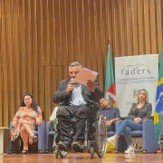 foto- no palco ao fundo o banner da faders, em cada lado uma bandeira ( do RS e do Brasil), a frente cadeiuras em couro azul onde estão sentados os representates dos 7 foruns regionais que aconteceram em 2024 e um representante dos Foruns de 2023, junto a coordenadora da Faders de Políticas Públicas, em primeiro plano o presidente da Faders em sua cadeira de rodas com micrfofone em punhos em uma das maos e na outra a carta aparesentada, faz seu pronunciamento. Fim da desscrição 