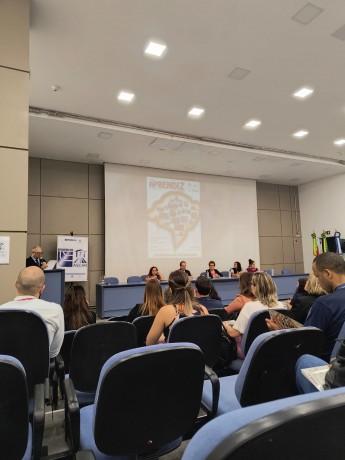 FOTO- AO FUNDO PALCO SENDO PROJETADA A CAPA DA REVISTA , NA FRENTE UMA MESA AZUL  COM CADEIRAS AZUIS ONDE ESTÃOS ENTADAS AS AUTORIDADES QUE PARTICIPARAM DO LANÇAMENTO DA REVISTA, AO LADO ESQUERDO UM PULPITO ONDE UM SENHOR DE CABELOS GRISALGOS, OCULOS E TERNO PRETO FAZ O CERIMONIAL, AO SEU LADO MAIS AO FUNDO UM BANNER DA EMATER RS. NA FRENTE DIVERSAS CADEIRAS COM ENCOSTOS AZUIS ONDE ESTA O PUBLICO. FIM DA DESCRIÇÃO