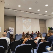 FOTO- AO FUNDO PALCO SENDO PROJETADA A CAPA DA REVISTA , NA FRENTE UMA MESA AZUL  COM CADEIRAS AZUIS ONDE ESTÃOS ENTADAS AS AUTORIDADES QUE PARTICIPARAM DO LANÇAMENTO DA REVISTA, AO LADO ESQUERDO UM PULPITO ONDE UM SENHOR DE CABELOS GRISALGOS, OCULOS E TERNO PRETO FAZ O CERIMONIAL, AO SEU LADO MAIS AO FUNDO UM BANNER DA EMATER RS. NA FRENTE DIVERSAS CADEIRAS COM ENCOSTOS AZUIS ONDE ESTA O PUBLICO. FIM DA DESCRIÇÃO