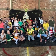 FOTO- no ginásio do Cazon, no zenário que foi montado para a apresentação circense, estão os artistas, usuários, familiares, e funcionários do Cazon, alfuns em pé, outros sentados, todos muito felizes e sorrindo, no fundo tem um pano preto e no chão também e ainda ao fundo dá pra ver um pinheiro de natal iluminado. Fim da descrição