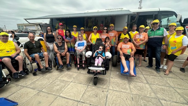 Foto no calçadão na beira da praia de Cidreira, ao fundo o ônibus da Faders- Rede Praia Acessível, na frente em primeiro plano a equipe da Faders que atua no programa e o diretor presidente Marco Antonio Lang, o Vice-Governador do RS Gabriel Souza, ao lado de sua esposa e filha.... o diretor do Procon e salva-vidas do corpo de Bombeiros Militar do RS.... ao centro a aluna da Apae de Feliz, Betina Wappler na cadeira anfíbia e que participou do primeiro banho assistido. Ao seu lado demais cadeirantes que estava presente visitando o Onibus.