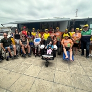 Foto no calçadão na beira da praia de Cidreira, ao fundo o ônibus da Faders- Rede Praia Acessível, na frente em primeiro plano a equipe da Faders que atua no programa e o diretor presidente Marco Antonio Lang, o Vice-Governador do RS Gabriel Souza, ao lado de sua esposa e filha.... o diretor do Procon e salva-vidas do corpo de Bombeiros Militar do RS.... ao centro a aluna da Apae de Feliz, Betina Wappler na cadeira anfíbia e que participou do primeiro banho assistido. Ao seu lado demais cadeirantes que estava presente visitando o Onibus.