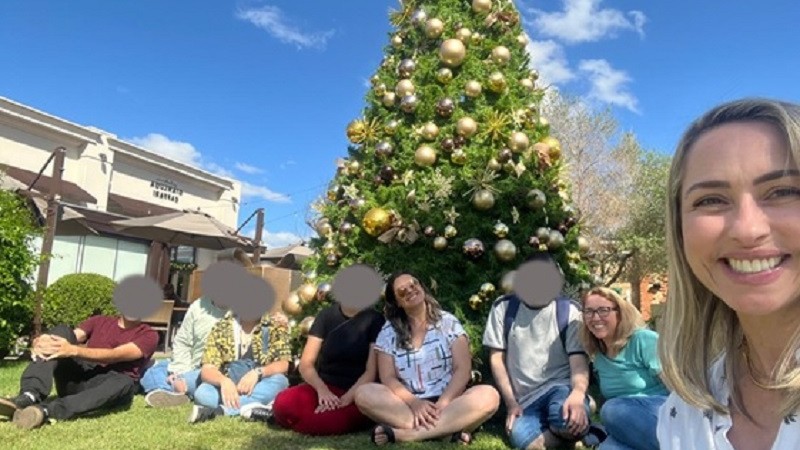 foto - ao fundo um prédio bege, e um pinheiro de natal grande natural e todo enfeitado em tons de dourado, em primeiro plano sentados em um gramado os usuários do Grupo Conexões Atípicas, e as coordenadoras do projeto, uma delas esta ebm na frente, pq é um selfie. Fim da descrição.