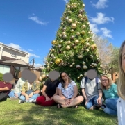 foto - ao fundo um prédio bege, e um pinheiro de natal grande natural e todo enfeitado em tons de dourado, em primeiro plano sentados em um gramado os usuários do Grupo Conexões Atípicas, e as coordenadoras do projeto, uma delas esta ebm na frente, pq é um selfie. Fim da descrição.