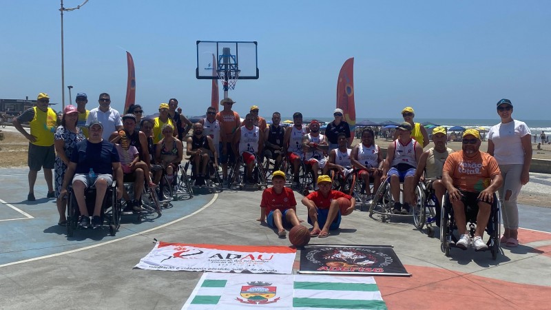 Foto: cadeirantes que participaram das atividades posam para foto em uma quadra de basquete, um ao lado do outro, ao fundo o mar, e a frente as bandeiras do municípios e da ADAU. Fim da descrição