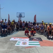 Foto: cadeirantes que participaram das atividades posam para foto em uma quadra de basquete, um ao lado do outro, ao fundo o mar, e a frente as bandeiras do municípios e da ADAU. Fim da descrição