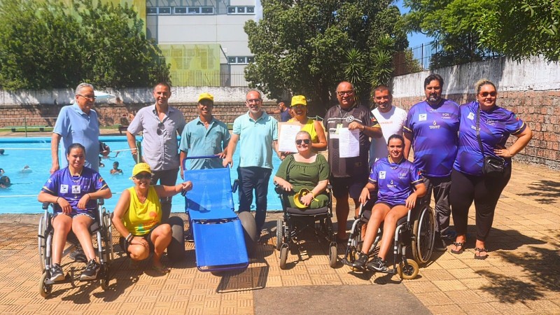 Foto - no fundo a piscina, na frente em primeiro plano, a cadeira anfíbia, as parateltas e autoridades que párticiparam da abertura dos banhos assistidos através do Programa Rede Praia Acessível da Faders. Fim da Descrição