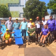 Foto - no fundo a piscina, na frente em primeiro plano, a cadeira anfíbia, as parateltas e autoridades que párticiparam da abertura dos banhos assistidos através do Programa Rede Praia Acessível da Faders. Fim da Descrição