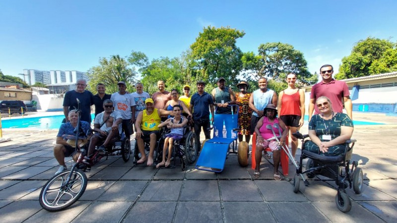 Foto: ao fundo da Piscina do Cevi, a frente em primeiro planom o presidente da Faders ao lado da cadeira anfíbia e demais cadeirantes que estavam no evento, em pés atrás deles as demais autoridades e famílias que participaram da entrega da cadeira que agora será utlizada para banhos assistidos para pessoas com deficiência e pessoas com mobilidade reduzida. Fim da descrição